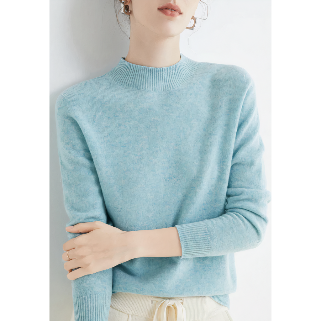Femme portant un pull bleu clair en cachemire, manches longues, col rond, posant contre un mur blanc. Mode féminine élégante et confortable.