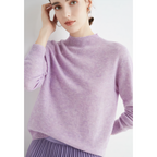 Femme portant un pull en cachemire lavande, manches longues, col rond, jupe plissée violette, posant contre un mur blanc. Mode élégante et tendance.