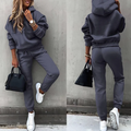 Femme en survêtement gris, baskets blanches, sac à main noir, posant contre un mur gris. Mode décontractée, tenue confortable, style urbain.