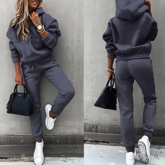 Femme en survêtement gris, baskets blanches, sac à main noir, posant contre un mur gris. Mode décontractée, tenue confortable, style urbain.