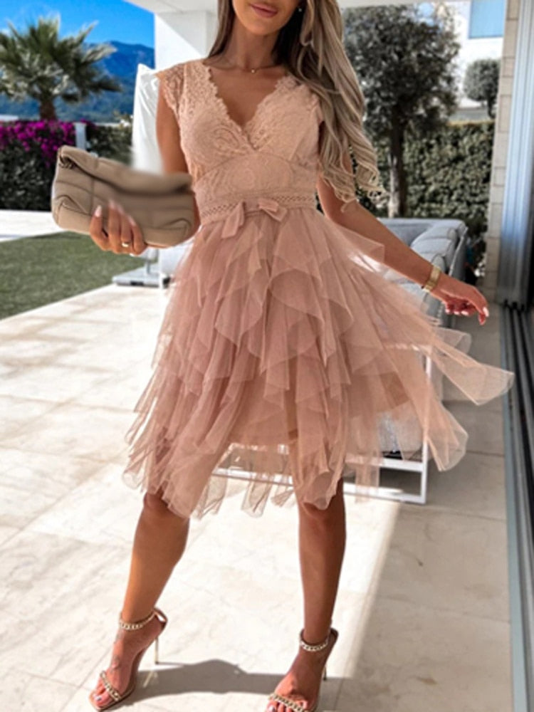 Robe rose en dentelle élégante, femme tenant une pochette beige, debout sur une terrasse ensoleillée avec vue sur jardin et montagnes.