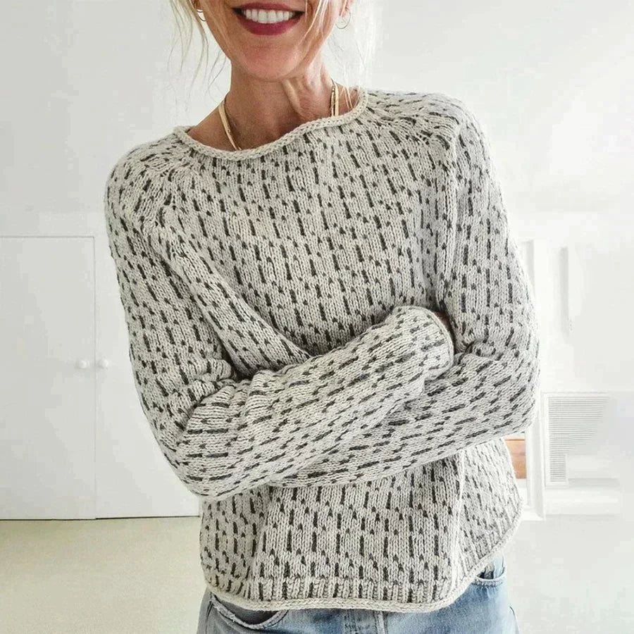 Femme souriante portant un pull en tricot gris à motifs, bras croisés, dans un intérieur lumineux. Mode automne-hiver, confort et style décontracté.
