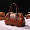 Sac à main en cuir marron élégant avec poignées robustes, fermetures éclair argentées et plaque métallique, posé sur une table en tissu beige.
