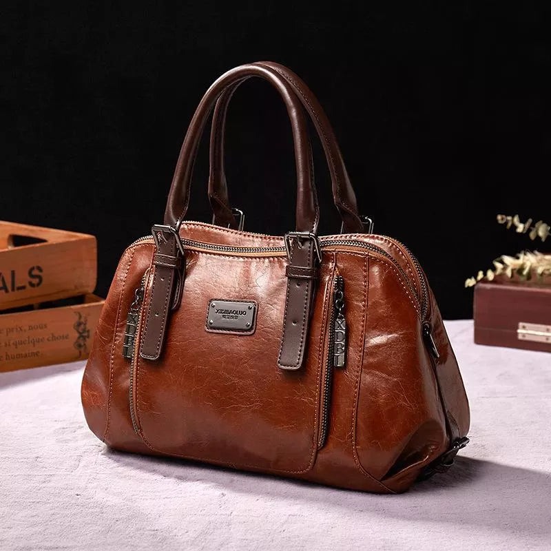 Sac à main en cuir marron élégant avec poignées robustes, fermetures éclair argentées et plaque métallique, posé sur une table en tissu beige.