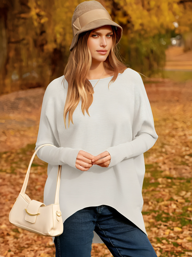 Femme en pull blanc et chapeau beige, portant un sac à main crème, posant dans un parc automnal avec feuilles jaunes. Mode automne, style décontracté.