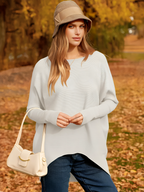 Femme en pull blanc et chapeau beige, portant un sac à main crème, posant dans un parc automnal avec feuilles jaunes. Mode automne, style décontracté.