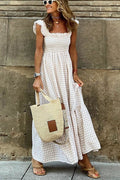 Femme en robe longue à carreaux beige, portant un sac en paille et des sandales, posant devant un mur en pierre. Mode estivale chic et décontractée.