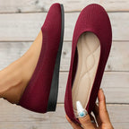 Ballerines rouges élégantes en tissu texturé, semelle noire, tenues par une main avec bague turquoise, posées sur fond de bois clair.