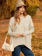 Femme en pull beige et chapeau, posant dans un parc automnal avec feuilles jaunes. Mode automne, style décontracté, sac à main blanc.