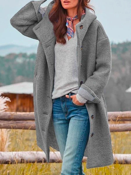 Femme portant un manteau gris oversize, pull gris, jean bleu, foulard coloré, posant devant une clôture en bois avec paysage naturel en arrière-plan.