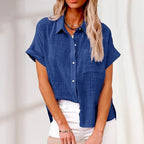 Chemisier bleu en lin pour femme, manches courtes, col classique, boutons blancs, porté avec un pantalon blanc. Mode féminine élégante et décontractée.