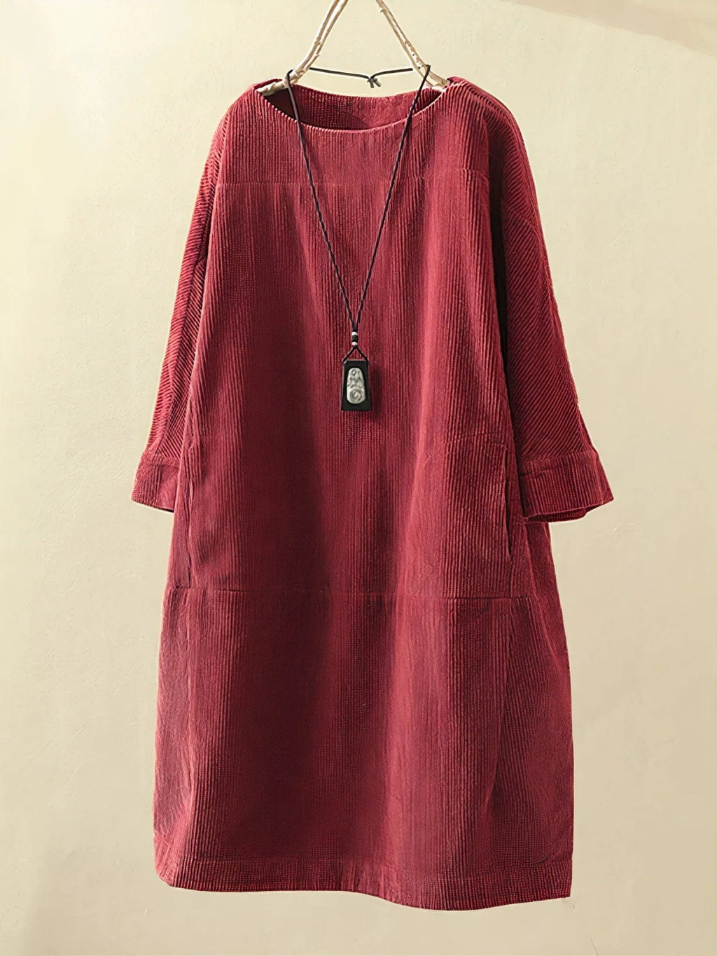 Robe en velours côtelé rouge bordeaux avec manches longues, suspendue sur un cintre, accompagnée d'un collier noir. Mode automne-hiver élégante.