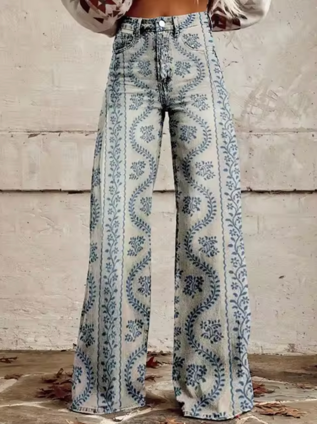 Jean évasé taille haute avec motif floral bleu sur fond beige, style vintage bohème, mode femme automne, devant mur texturé.