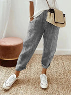 Personne portant un pantalon rayé bleu et blanc, t-shirt blanc, baskets blanches, sac en osier, debout sur un tapis beige. Mode décontractée chic.