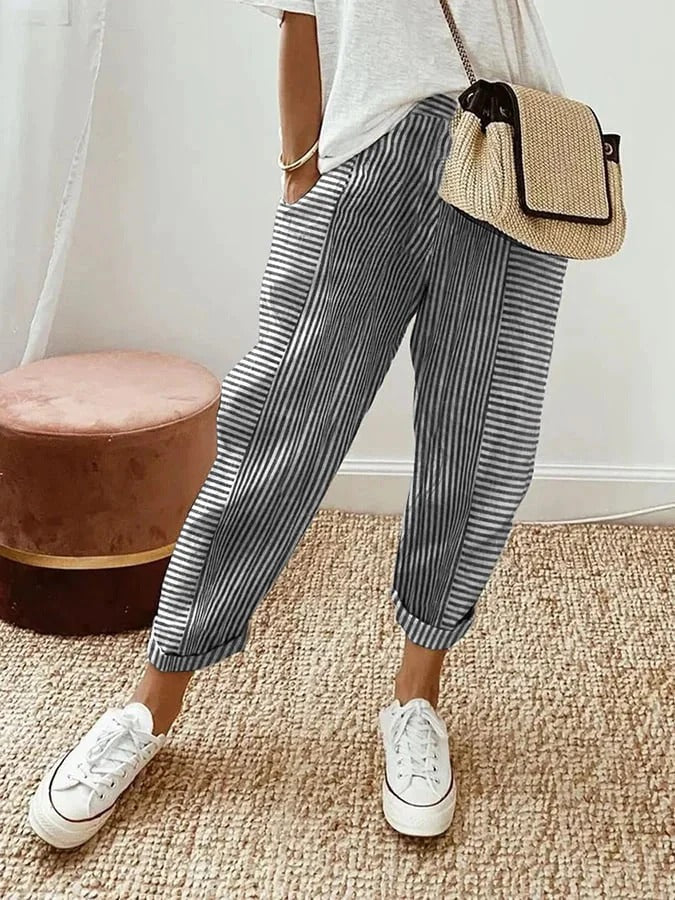 Personne portant un pantalon rayé bleu et blanc, t-shirt blanc, baskets blanches, sac en osier, debout sur un tapis beige. Mode décontractée chic.