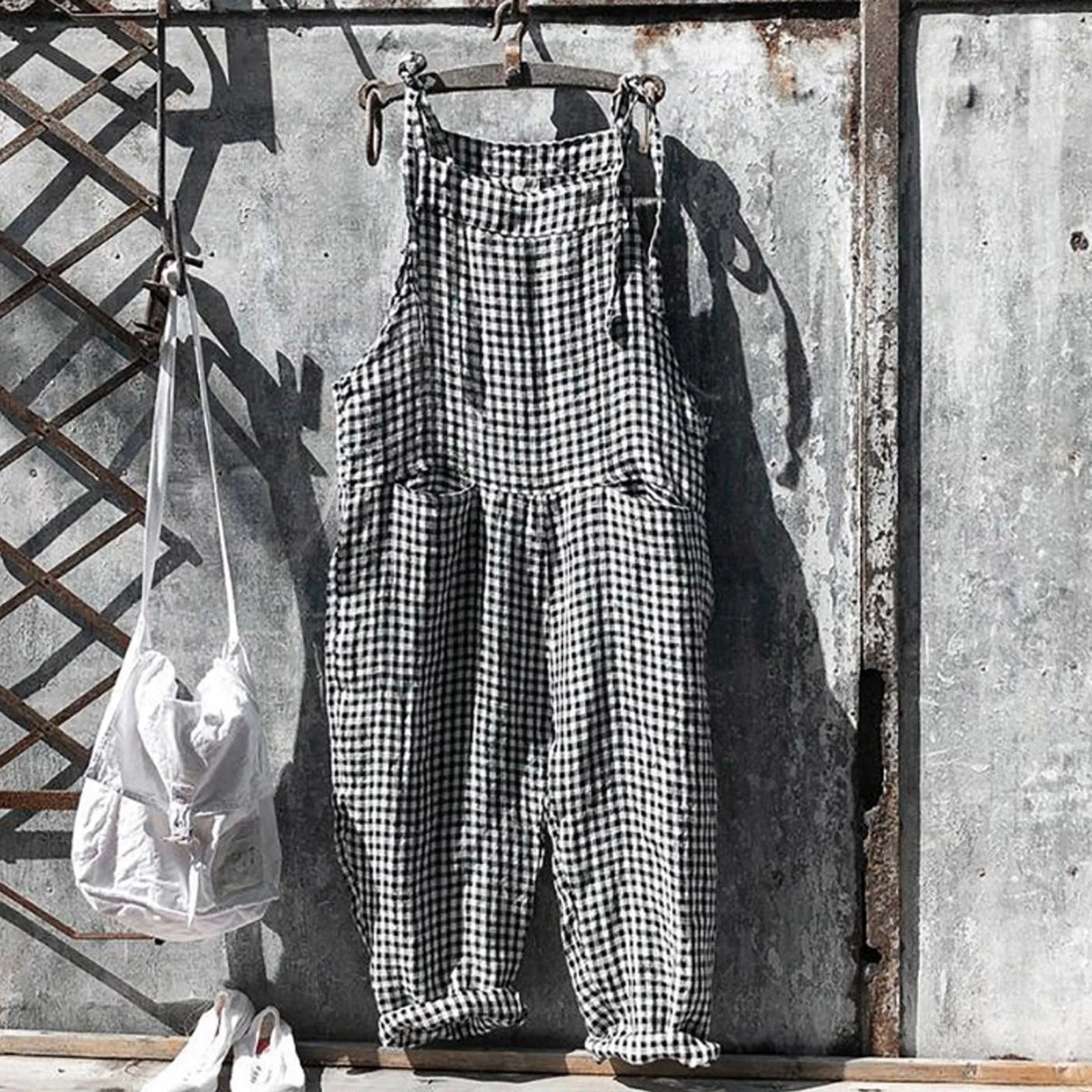 Salopette à carreaux noir et blanc suspendue sur un mur en métal rouillé, sac blanc à côté. Mode vintage, style rétro, vêtements décontractés.