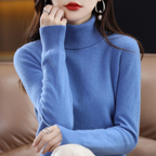 Femme portant un pull bleu en cachemire, col roulé, assise sur un canapé en cuir. Accessoires: boucles d'oreilles, bague. Mode élégante automne-hiver.