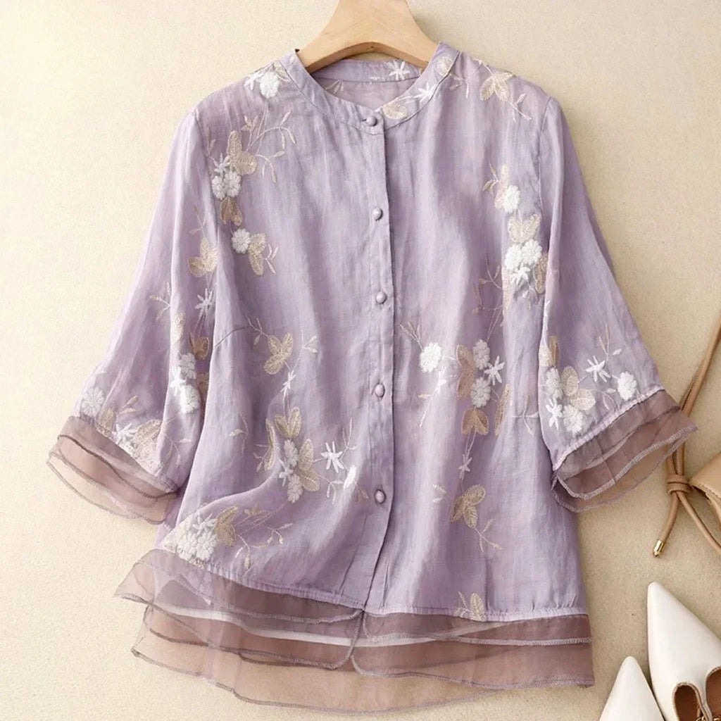 Blouse en lin violet clair avec broderie florale blanche et beige, manches trois-quarts, col rond, boutons, style élégant et féminin.