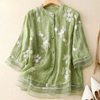 Blouse verte en lin avec broderie florale blanche, manches trois-quarts, col rond, boutons assortis, posée sur un cintre en bois.