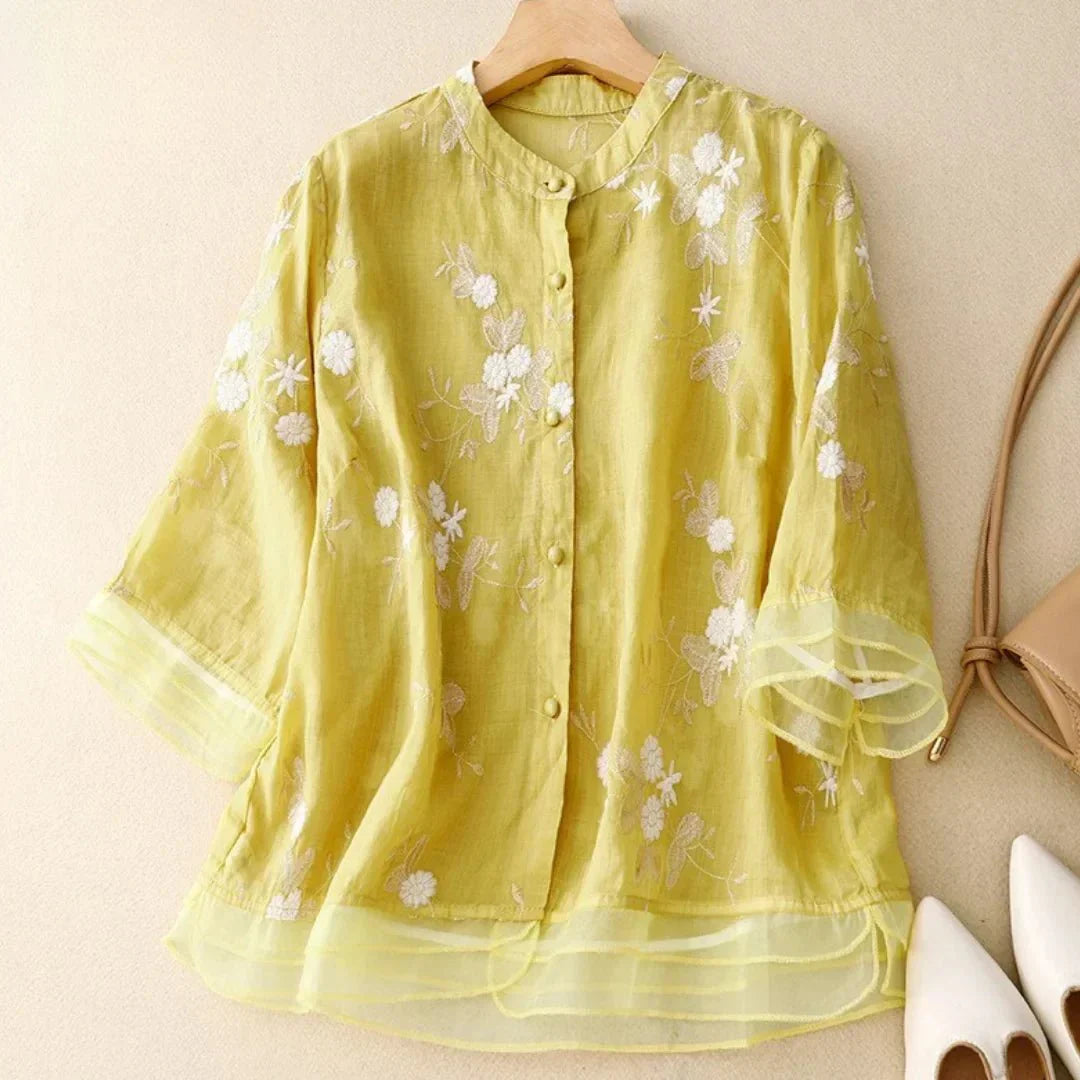 Blouse en lin jaune avec broderie florale blanche, manches trois-quarts, col rond, posée sur un cintre, accompagnée de chaussures blanches et sac beige.