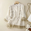 Blouse en mousseline blanche à motifs floraux, manches trois-quarts, posée sur un cintre en bois, avec sac à chaîne dorée et sandales blanches à côté.