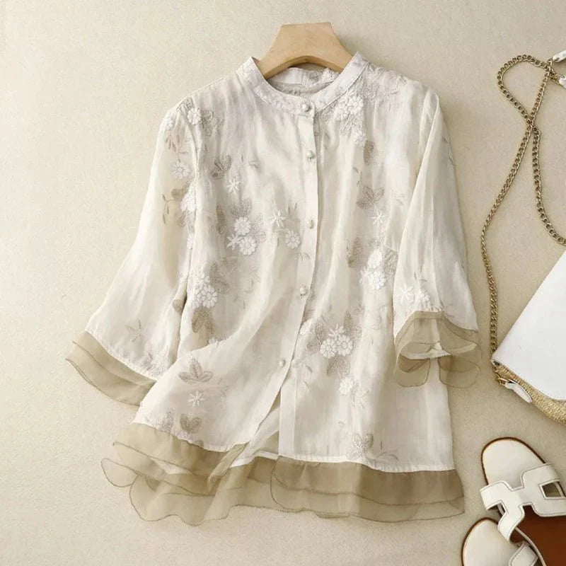 Blouse en mousseline blanche à motifs floraux, manches trois-quarts, posée sur un cintre en bois, avec sac à chaîne dorée et sandales blanches à côté.