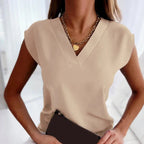 Femme portant un haut beige élégant avec col en V, collier chaîne doré avec pendentif cœur, tenant une pochette noire. Mode féminine chic et moderne.