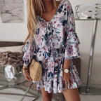 Robe à fleurs pastel avec manches à volants, portée par une femme blonde. Accessoires: montre dorée, sac en osier. Décor intérieur moderne.