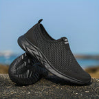 Chaussure de sport noire en maille respirante sur fond de plage, semelle antidérapante, idéale pour la course et les activités extérieures.