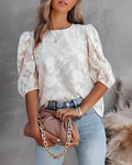 Femme en blouse blanche à motifs floraux, manches bouffantes, tenant un sac à main beige avec chaîne dorée, portant un jean bleu clair.