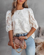 Femme en blouse blanche à motifs floraux, manches bouffantes, tenant un sac à main beige avec chaîne dorée, portant un jean bleu clair.