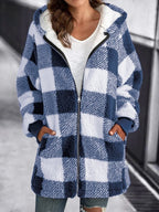 Femme portant un manteau à carreaux bleu et blanc, style décontracté, avec capuche et poches, sur fond urbain. Mode automne-hiver tendance.