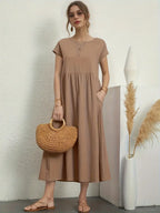 Femme en robe longue beige, tenant un sac en osier, posant près d'un vase blanc avec des pampas. Mode élégante, style décontracté chic.