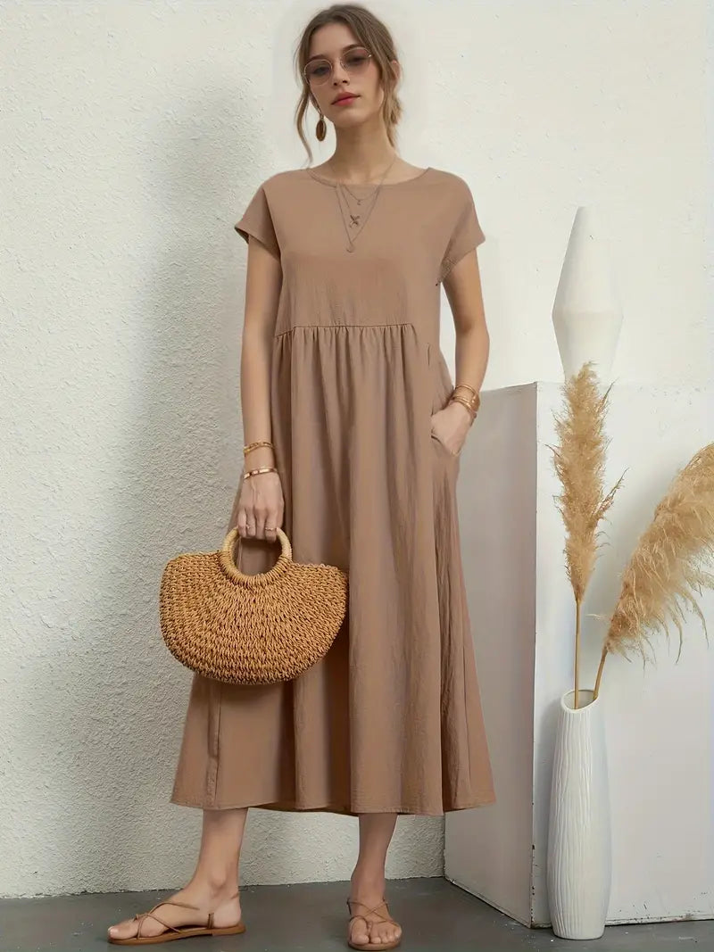 Femme en robe longue beige, tenant un sac en osier, posant près d'un vase blanc avec des pampas. Mode élégante, style décontracté chic.