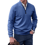 Homme portant un pull bleu à col zippé, chemise blanche et jean, tenant une veste marron. Mode masculine élégante et décontractée.