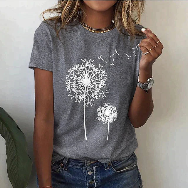 Femme portant un t-shirt gris avec motif pissenlit blanc, jean décontracté, montre argentée, collier doré, posant devant un mur blanc.