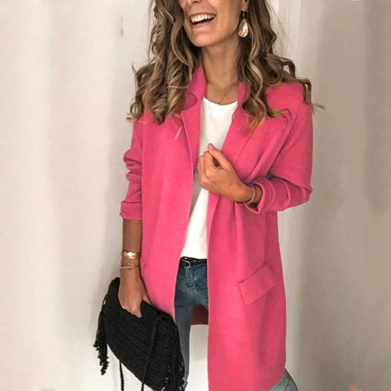 Femme souriante portant un manteau rose vif, chemise blanche et jeans, tenant un sac noir. Mode féminine élégante et moderne.