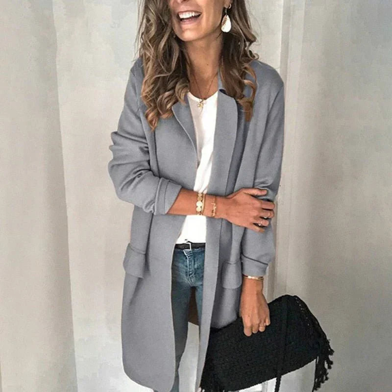 Femme souriante portant un manteau gris élégant, jeans décontracté, sac noir à franges, style automne chic, mode féminine tendance.