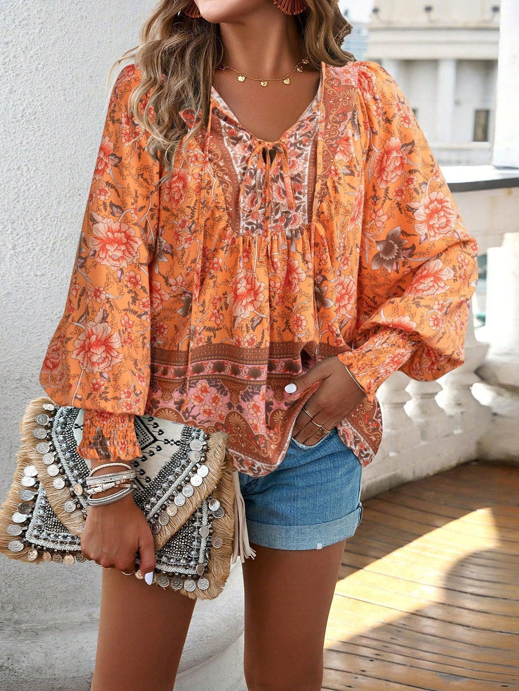 Femme portant un chemisier bohème orange à motifs floraux, short en jean, et tenant un sac à main en paille décoré de perles. Mode estivale chic.