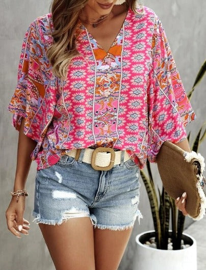 Femme portant un haut bohème rose à motifs géométriques et short en jean déchiré, accessoirisée d'une ceinture beige et d'un sac en paille.