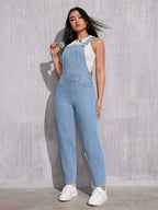 Femme portant une salopette en jean bleu clair, posant contre un mur gris. Mode décontractée, tendance, style urbain, chaussures blanches.