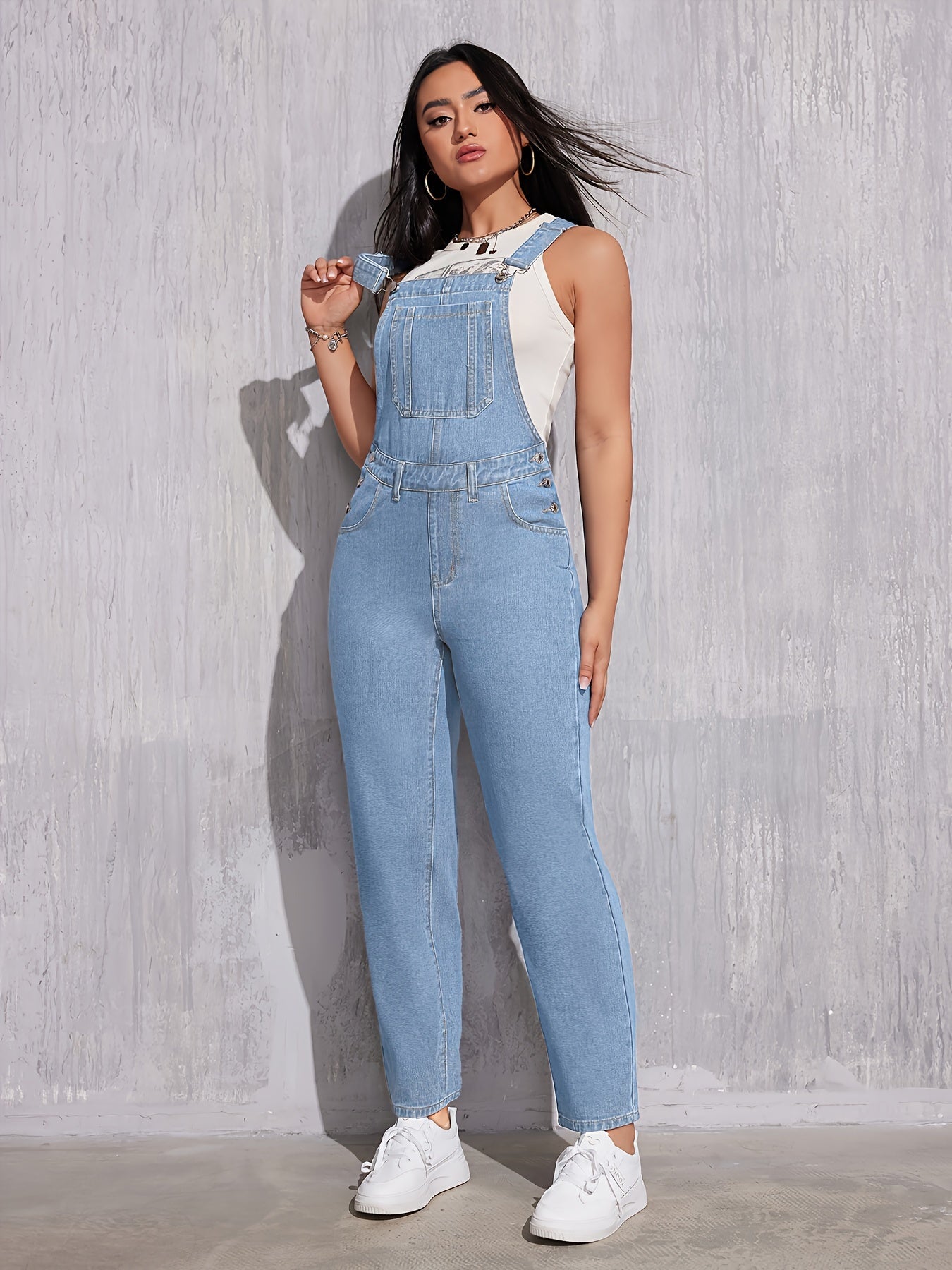Femme portant une salopette en jean bleu clair, posant contre un mur gris. Mode décontractée, tendance, style urbain, chaussures blanches.