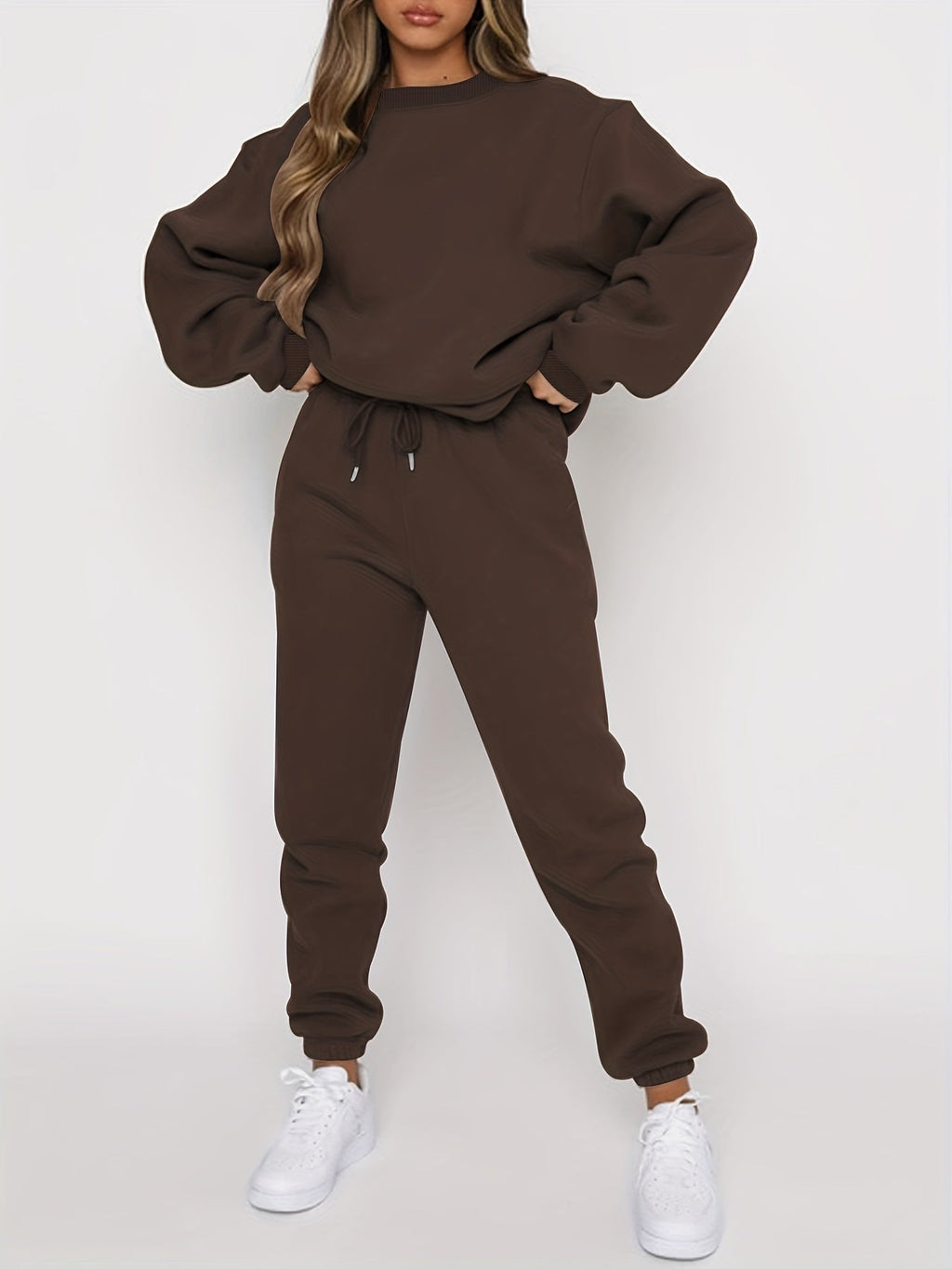 Femme portant un ensemble de survêtement marron, composé d'un sweat ample et d'un pantalon de jogging, avec des baskets blanches. Mode décontractée tendance.