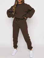 Femme portant un ensemble de survêtement marron, composé d'un sweat ample et d'un pantalon de jogging, avec des baskets blanches. Mode décontractée tendance.