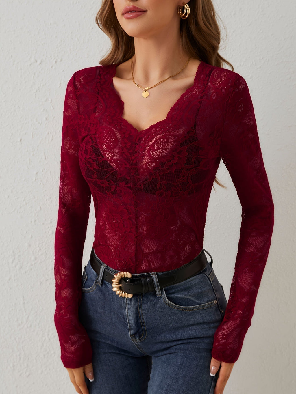 Femme portant un haut en dentelle rouge bordeaux à manches longues, col en V, avec un jean bleu et une ceinture noire. Mode élégante et tendance.