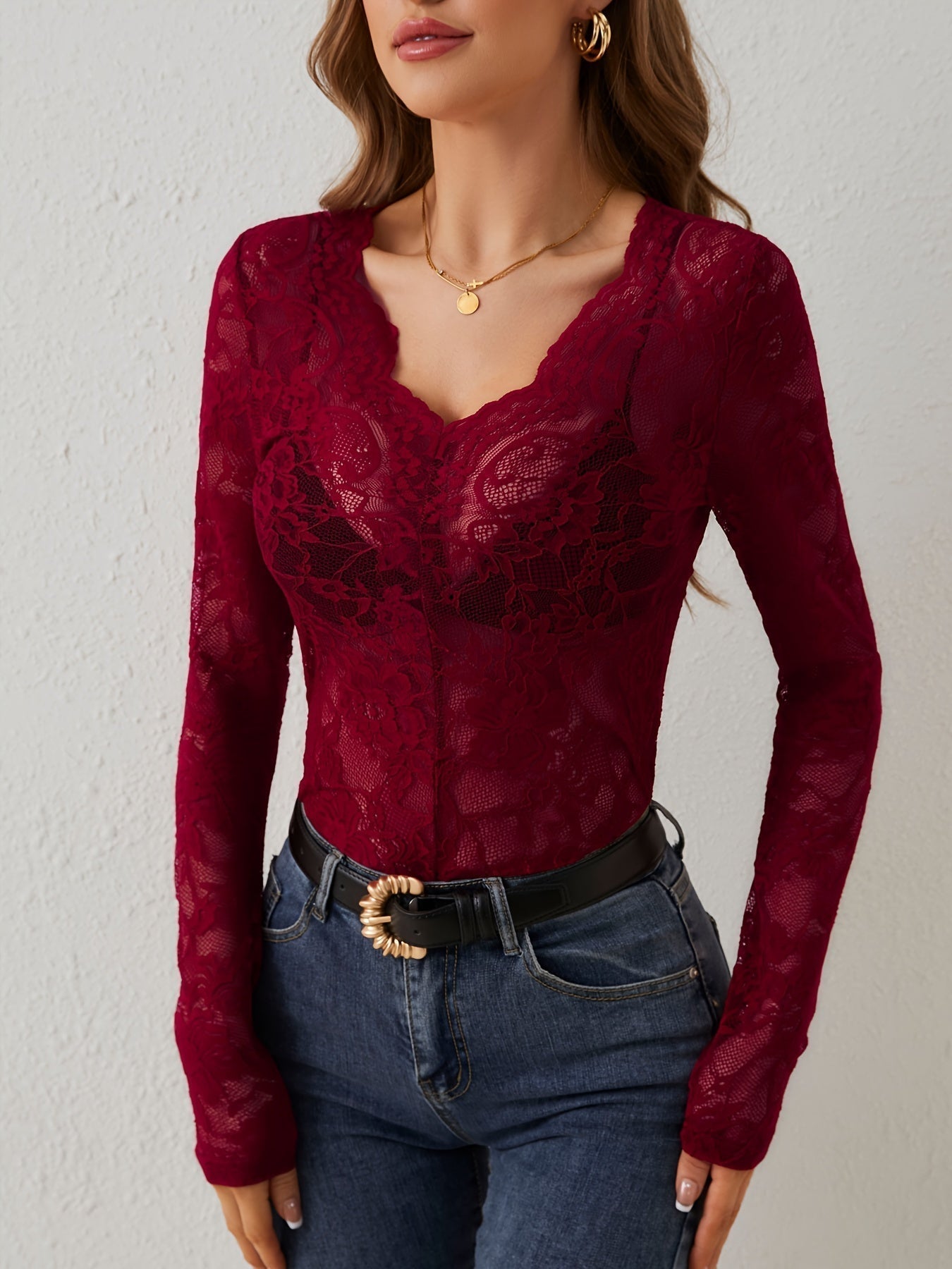 Femme portant un haut en dentelle rouge bordeaux à manches longues, col en V, avec un jean bleu et une ceinture noire. Mode élégante et tendance.