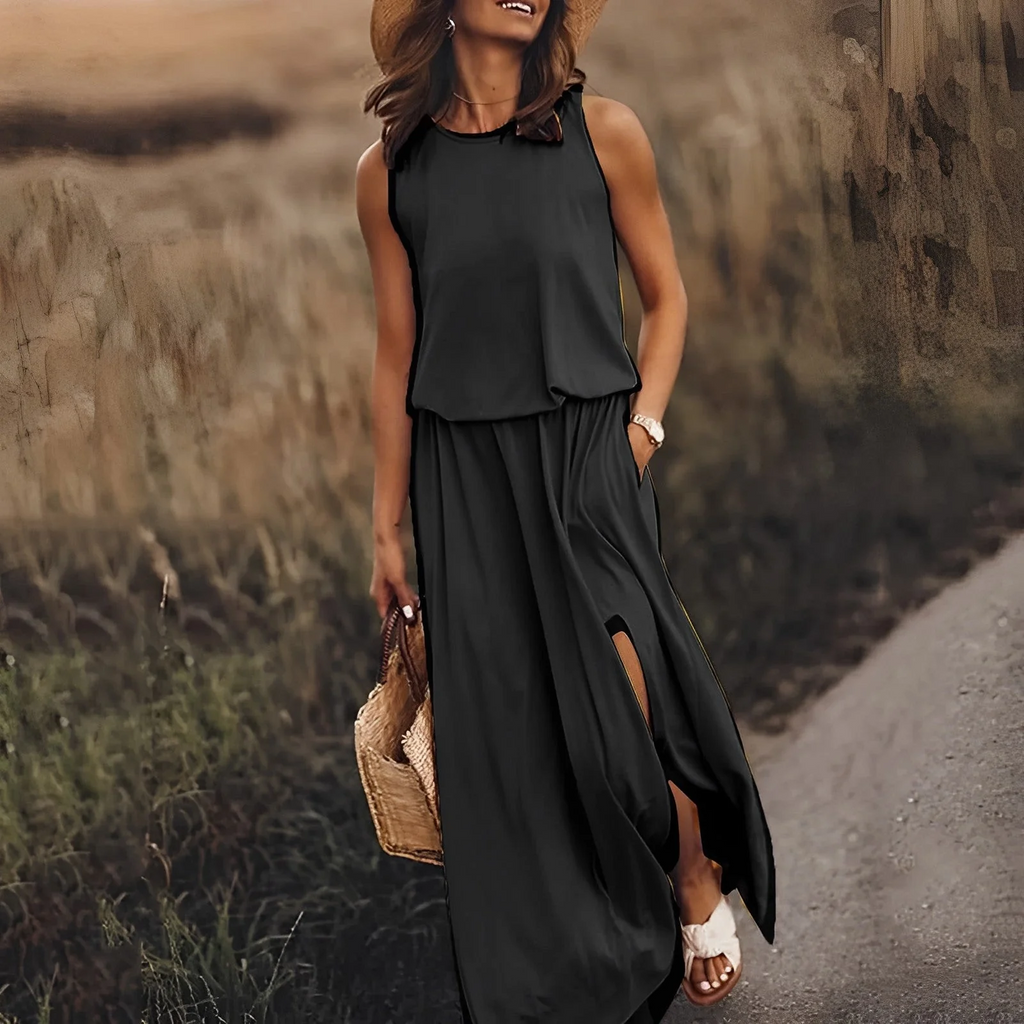 Femme en robe longue noire, chapeau de paille, sandales blanches, tenant un sac en osier, marchant dans un champ au coucher du soleil. Mode été chic.