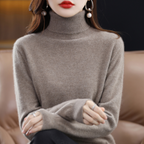 Femme portant un pull col roulé en cachemire beige, boucles d'oreilles pendantes, lèvres rouges, assise sur un canapé en cuir. Mode hiver élégante.