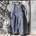 Salopette à carreaux bleus et blancs suspendue à un mur en métal vieilli, accompagnée d'un sac blanc et de chaussures blanches. Mode vintage décontractée.