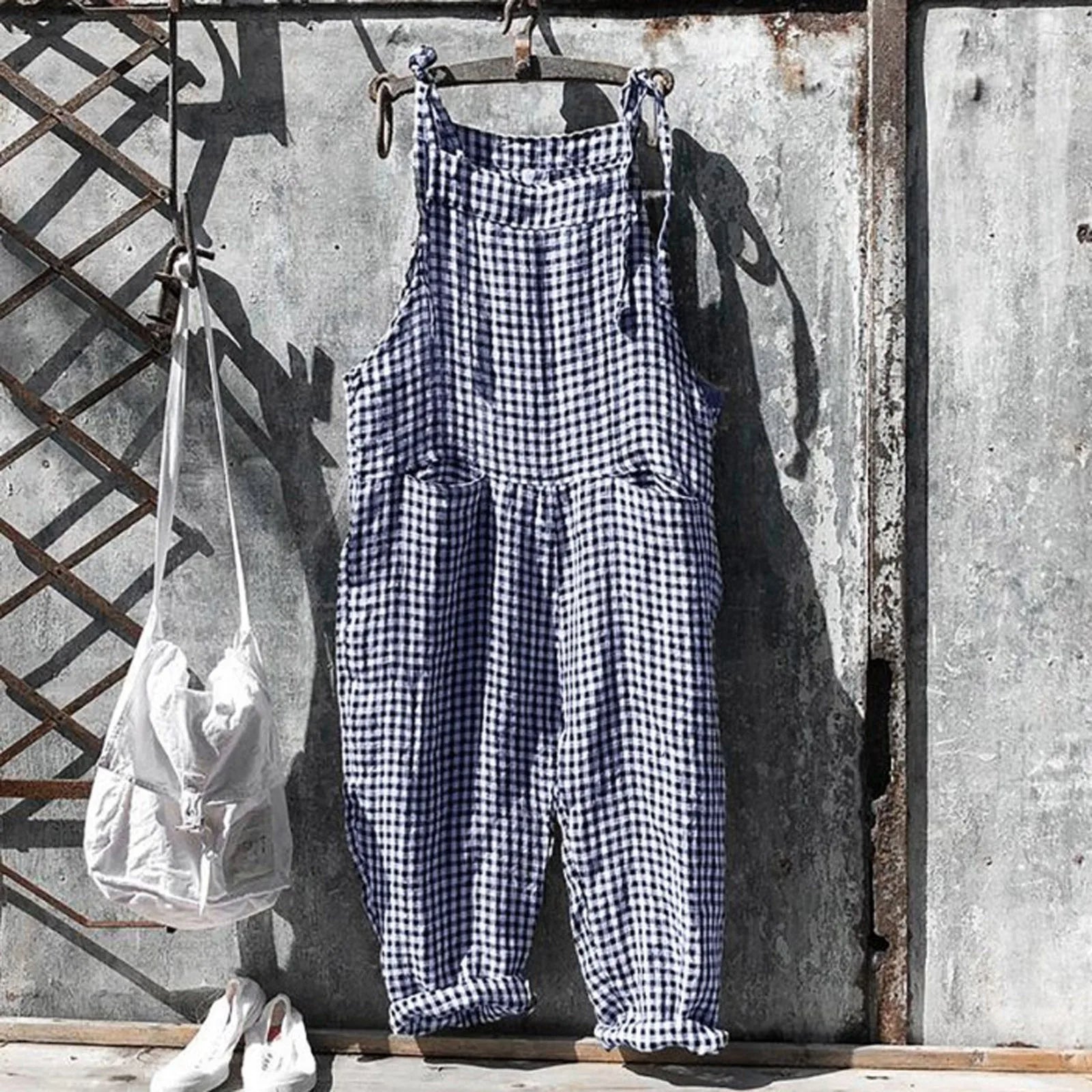 Salopette à carreaux bleus et blancs suspendue à un mur en métal vieilli, accompagnée d'un sac blanc et de chaussures blanches. Mode vintage décontractée.
